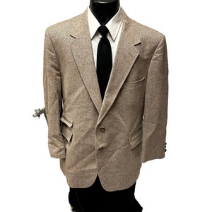 Puede incluir: Una chaqueta de tweed marrón con cierre de dos botones y bolsillos de solapa. La chaqueta tiene un sutil patrón de espiga y se lleva con una corbata negra.