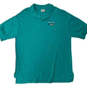 Op de afbeelding: Turquoise polo shirt met de tekst "The Queen's Hotel Casino Las Vegas" geborduurd op de linkerborst.