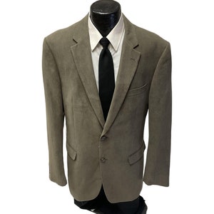 May include: A brown suede blazer with a two-button closure and a single chest pocket. The blazer has a white collared shirt underneath and a black tie.