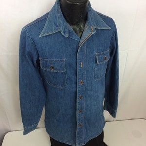 Puede incluir: Una camisa de mezclilla azul de manga larga con cierre de botones y dos bolsillos de solapa. La camisa tiene un cuello puntiagudo y un pu&ntilde;o de un bot&oacute;n.