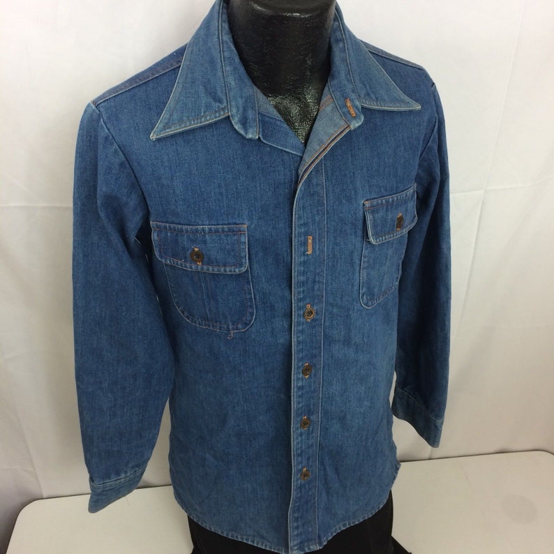 Puede incluir: Una camisa de mezclilla azul de manga larga con cierre de botones y dos bolsillos de solapa. La camisa tiene un cuello puntiagudo y un pu&ntilde;o de un bot&oacute;n.