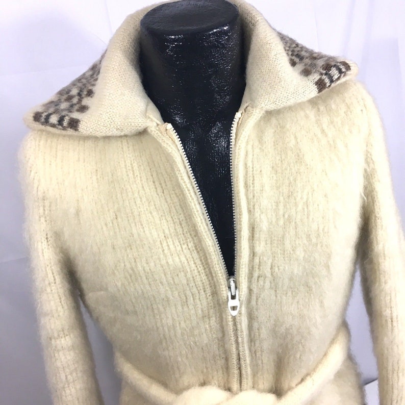 K&ouml;nnte beinhalten: Ein wei&szlig;er, flauschiger Cardigan mit Rei&szlig;verschluss, einem braunen und wei&szlig;en Muster am Kragen und einem passenden G&uuml;rtel.