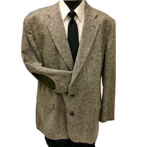 Puede incluir: Una blazer de tweed gris con coderas verde oscuro, una camisa blanca y una corbata negra. La blazer tiene un cierre clásico de dos botones y solapas con muescas. El aspecto general es profesional y adecuado para negocios u ocasiones formales.