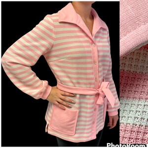 Puede incluir: Una camisa de manga larga a rayas rosa y blancas con cuello rosa y cinturón a juego. La camisa tiene cierre de botones en la parte delantera y un bolsillo de parche en el lado izquierdo.