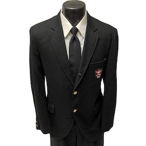 May include: A black blazer with a gold button closure and a patch on the pocket. The blazer has a white collared shirt underneath with a black tie.