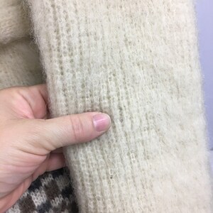 K&ouml;nnte beinhalten: Nahaufnahme eines cremefarbenen Strickstoffes mit einer flauschigen Textur. Der Stoff ist weich und flauschig, mit einem dezenten Rippenmuster.