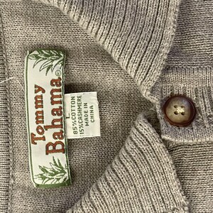 May include: A gray Tommy Bahama sweater with a button closure and a label that reads "Tommy Bahama" and "85% Cotton 15% Cashmere Made in China".