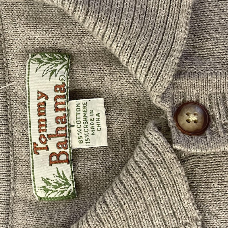 May include: A gray Tommy Bahama sweater with a button closure and a label that reads "Tommy Bahama" and "85% Cotton 15% Cashmere Made in China".
