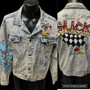 May include: A light blue denim jacket with graffiti art on the front and back. The front features a blue and yellow graphic of the word "Slick" and a yellow circle with the letter "S" inside. The back features a black and white chequered pattern, red and yellow graffiti, and the word "Slick" in red, yellow, and orange.