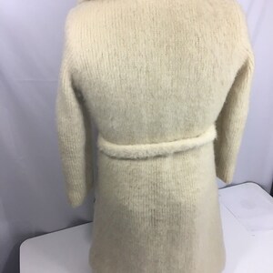 K&ouml;nnte beinhalten: Ein langer, wei&szlig;er, flauschiger Cardigan mit einem braunen und wei&szlig;en karierten Kragen und einem passenden G&uuml;rtel.