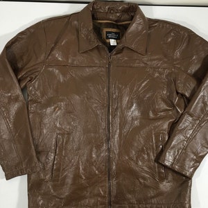 May include: A brown leather jacket with a zipper closure and a collar. The jacket has a pocket on the left side and a label that reads "Executive Division" on the inside of the collar.