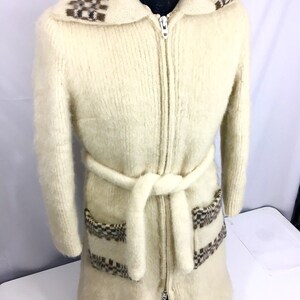 K&ouml;nnte beinhalten: Ein langer, wei&szlig;er, flauschiger Strickcardigan mit einem braunen und wei&szlig;en geometrischen Muster an Kragen, Taschen und G&uuml;rtel. Der Cardigan hat einen Rei&szlig;verschluss und einen G&uuml;rtel.