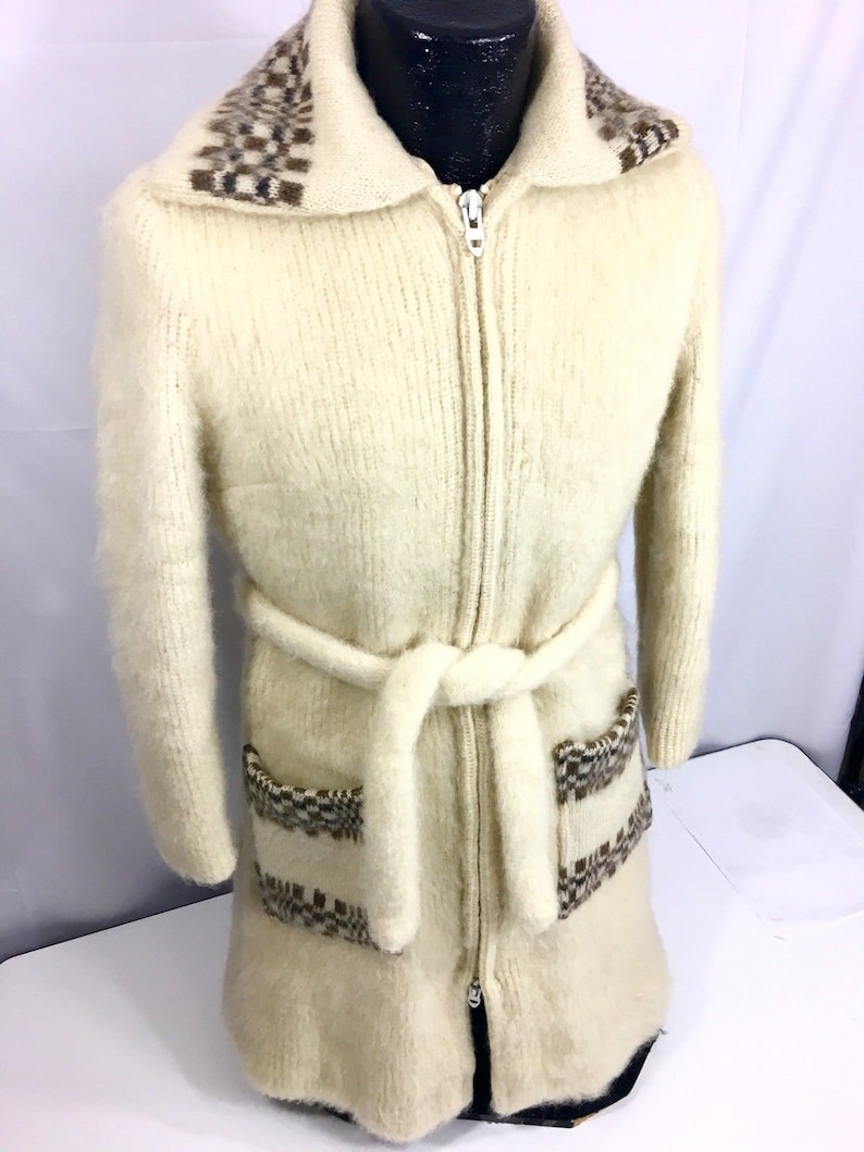 K&ouml;nnte beinhalten: Ein langer, wei&szlig;er, flauschiger Strickcardigan mit einem braunen und wei&szlig;en geometrischen Muster an Kragen, Taschen und G&uuml;rtel. Der Cardigan hat einen Rei&szlig;verschluss und einen G&uuml;rtel.
