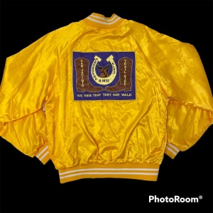 May include: Yellow satin bomber jacket with a purple patch on the back. The patch features a horseshoe, a deer, and the text "Pasadena Elks Trail Ride #1832 San Jacinto". The text "We Ride That They May Walk" is below the patch.