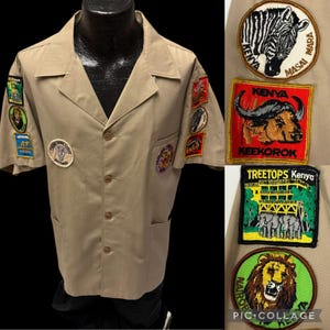 May include: A tan short-sleeved shirt with multiple embroidered patches featuring animals and locations from Kenya. The patches include a zebra, a buffalo, an elephant, and a lion. The shirt has a button-up front and two pockets.