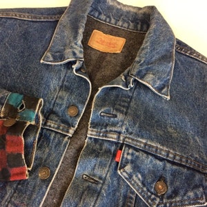 Op de afbeelding: Een blauwe denimjas met een rode en zwarte geruite stof die de manchetten bekleedt. De jas heeft een Levi's-label op de zak.