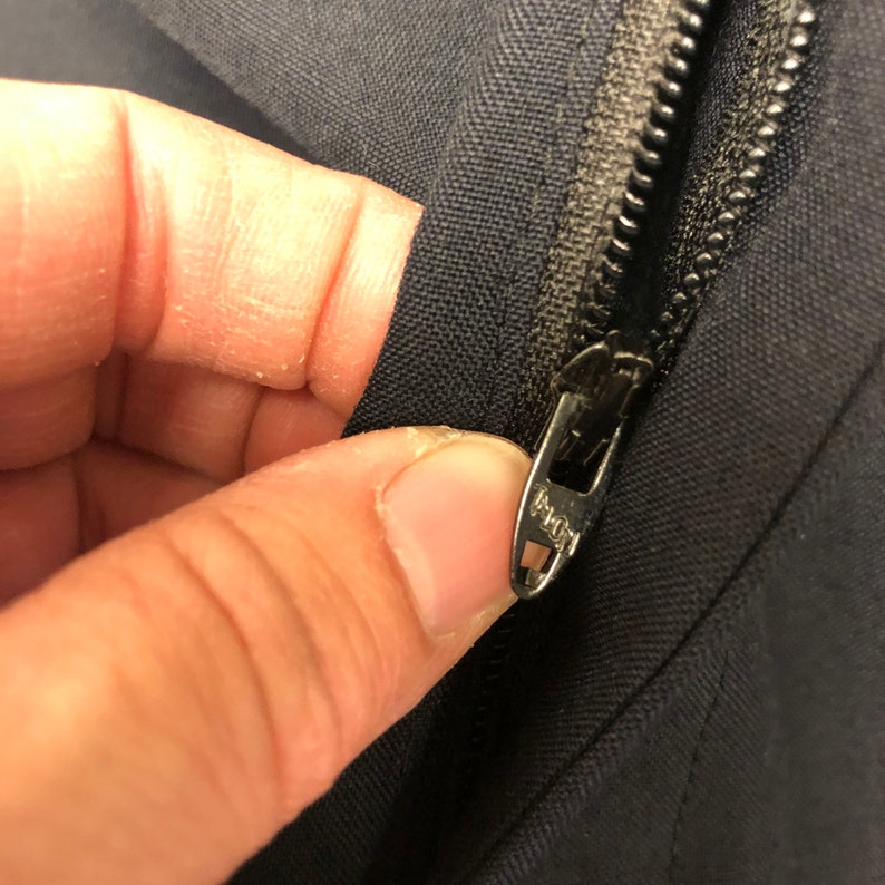 May include: Close-up of a black zipper with a metal slider. The zipper is partially open and the slider is pulled down. The zipper is sewn into a piece of black fabric.