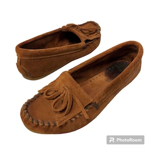 May include: A pair of brown suede moccasins with fringe detailing. The moccasins have a stitched seam around the edge and a leather sole.