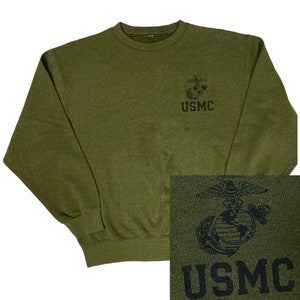 May include: A green sweatshirt with a black embroidered USMC logo on the front. The logo features an eagle, globe, and anchor.