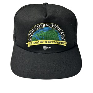 May include: Black baseball cap with a curved brim. The cap features a circular graphic with a globe design and the text "GOING GLOBAL WITH AT&T". Below the globe is the text "AT&T. FAR AND AWAY. THE BEST IN THE BUSINESS". The AT&T logo is below the text.