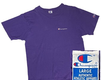 May include: Purple short-sleeved t-shirt with a white Champion logo on the front and a red and white Champion logo on the right sleeve. The tag says "Champion USA Large Authentic Athletic Apparel Made in U.S.A. Care on Reverse."