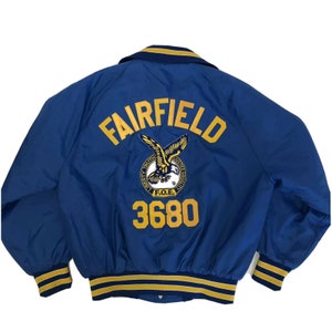 May include: Blue satin varsity jacket with yellow and white stripes on the sleeves and collar. The back of the jacket has the word "FAIRFIELD" in yellow letters with a yellow eagle logo and the number "3680" below it.
