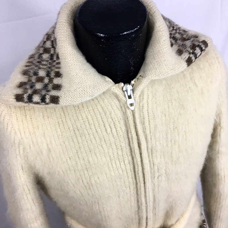 K&ouml;nnte beinhalten: Ein wei&szlig;er Strickcardigan mit einem braunen und wei&szlig;en gemusterten Kragen und einem wei&szlig;en Rei&szlig;verschluss. Der Cardigan hat einen G&uuml;rtel.