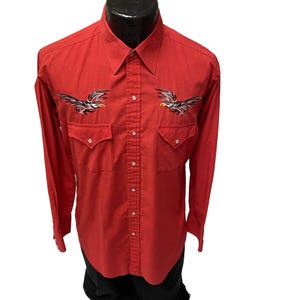 Puede incluir: Una camisa roja de manga larga con botones y águilas negras y blancas bordadas en el pecho.