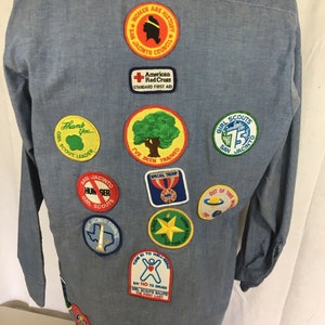 May include: A light blue denim shirt with various embroidered patches, including Girl Scout patches, a red cross patch, and a patch that says "Women Are History".