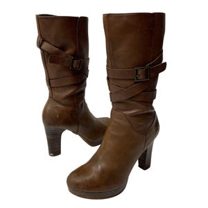 Puede incluir: Botas de cuero marrón con tacón alto y correas con hebilla.