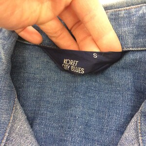 Puede incluir: Una camisa vaquera azul con una etiqueta azul que dice "Koret City Blues" y "S".
