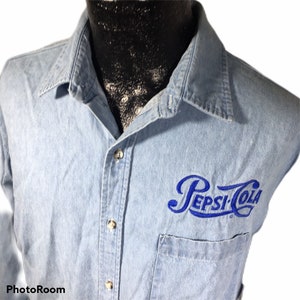 May include: Light blue denim shirt with the embroidered Pepsi-Cola logo in blue on the chest pocket. The shirt has a button-down front, a collar, and a single chest pocket. The shirt is a vintage style.