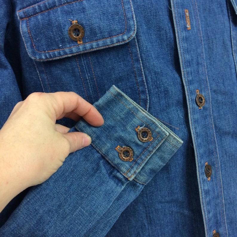 Puede incluir: Una camisa vaquera azul con cierre de botones en la parte delantera y un solo bolsillo en el pecho. La camisa tiene un pu&ntilde;o abotonado con la palabra "KORET" grabada en los botones.