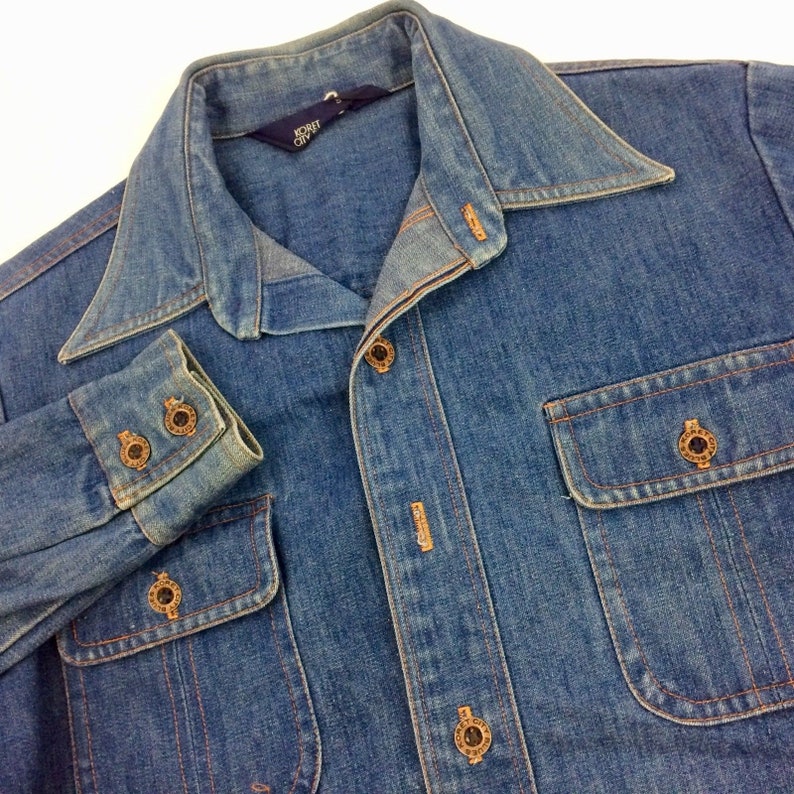 Puede incluir: Una camisa vaquera azul con cuello abotonado y mangas largas. La camisa tiene un bolsillo en el pecho izquierdo y dos bolsillos grandes en la parte inferior. La camisa tiene una etiqueta que dice "NORTH CITY".