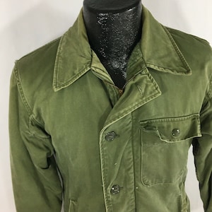 May include: A green military-style jacket with a zipper and button closure. The jacket has a large pocket on the front and a smaller pocket on the chest.