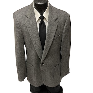 Puede incluir: Una chaqueta de tweed gris con una corbata negra y una camisa blanca de cuello. La chaqueta tiene dos bolsillos de parche y un cierre de botón.