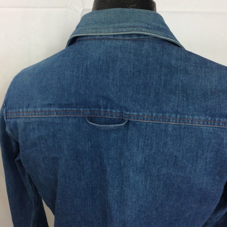 Puede incluir: Una camisa vaquera azul con un solo bolsillo trasero. La camisa tiene cuello abotonado y mangas largas.
