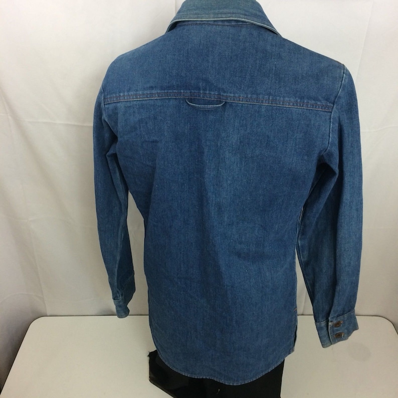 Puede incluir: Una camisa de mezclilla azul de manga larga con cuello abotonado y un solo bot&oacute;n en el pu&ntilde;o. La camisa tiene un yugo trasero y un solo bolsillo trasero.