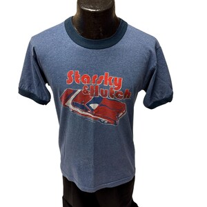 Könnte beinhalten: Blaues T-Shirt mit Rundhalsausschnitt und kontrastierenden Ärmeln, mit dem Text "Starsky & Hutch" in Rot und Weiß und einer roten und weißen Autografik.