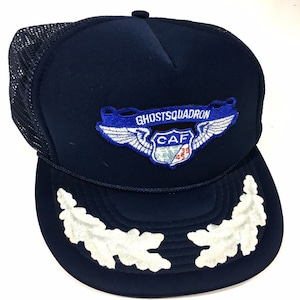 May include: A navy blue trucker hat with a white mesh back. The hat features a white embroidered patch with the text "Ghost Squadron" and a blue patch with the text "CAF 39 454". The hat also has two white embroidered oak leaves on the front.