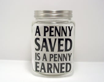 May include: A clear glass jar with a silver-colored metal lid. The front of the jar features black lettering that reads A PENNY SAVED IS A PENNY EARNED. The jar has a cylindrical shape with a wide mouth and a flat base, designed for storage.