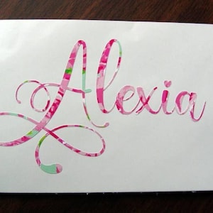 May include: A white decal with the name "Alexia" in a cursive font. The letters are filled with a pink, green, and white floral pattern. The decal is likely for a car, laptop, or other personal item.