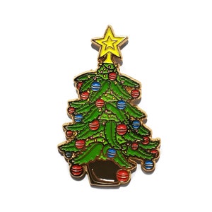 Puede incluir: Un pin de esmalte dorado de un árbol de Navidad verde con adornos rojos y azules, una estrella amarilla en la parte superior y una maceta marrón.