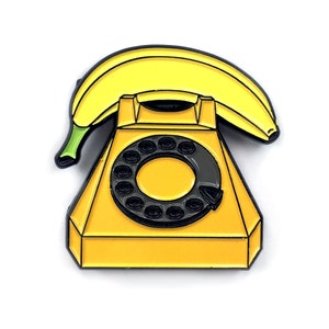 May include: A yellow enamel pin of a rotary phone with a banana for the receiver. The phone is yellow with a black dial and the banana is yellow with a brown stem.
