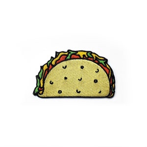 Puede incluir: Un parche bordado con forma de taco con una concha amarilla que presenta pequeños puntos negros. El relleno incluye capas de lechuga verde, tomate rojo y queso amarillo, todo delineado con un borde negro grueso sobre fondo blanco.