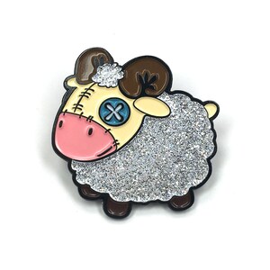 May include: A glittery silver and white enamel pin of a cartoon sheep with brown horns, a pink nose, and a blue button eye with an X on it.