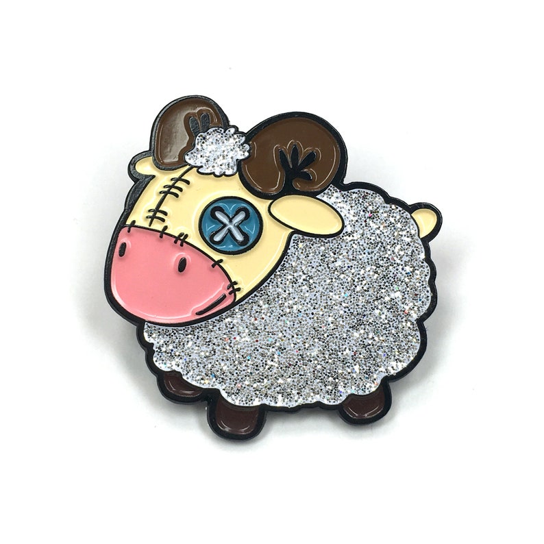 May include: A glittery silver and white enamel pin of a cartoon sheep with brown horns, a pink nose, and a blue button eye with an X on it.