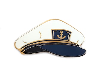 May include: A white and navy blue enamel pin shaped like a captain's hat. The design features a gold-colored metal outline, a dark blue brim, and a central navy blue rectangular patch displaying a gold anchor symbol on the front of the hat.