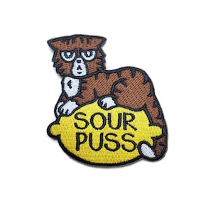 Puede incluir: Un parche bordado para planchar con un gato marrón con una expresión gruñona sentado en un limón amarillo con el texto "Sour Puss" en negro.