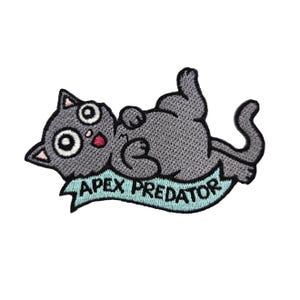 Puede incluir: Parche bordado para planchar con un gato de dibujos animados gris con ojos grandes y boca roja, tumbado de espaldas con las patas en el aire. El gato está sobre una pancarta azul claro con el texto "APEX PREDATOR" en negro.